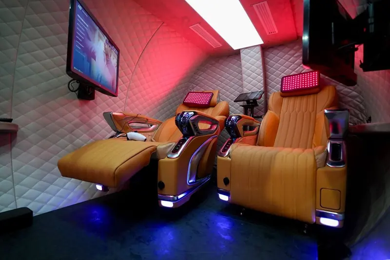 Lifetech interior: two tan leather recliners with red light panels at the head, a wall screen and a control tablet